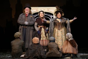  Three actors dressed as poor Victorian-era street vendors stand in front of a doll vendor’s cart telling a story to four seated children.