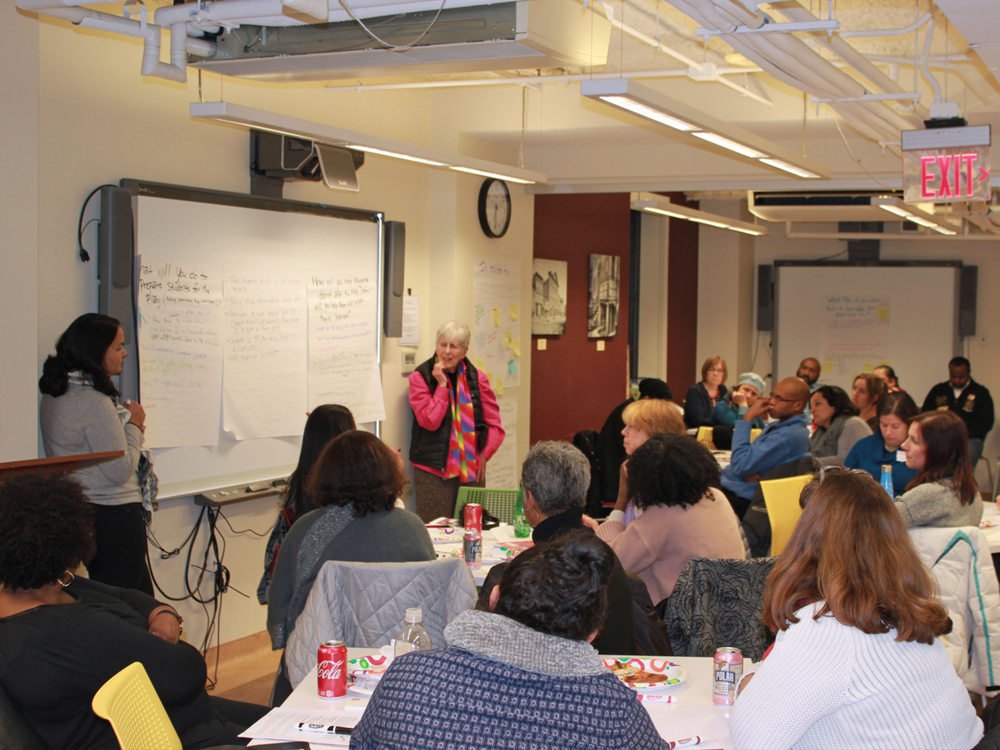 Teachers listen to a program facilitator who is speaking in front of a white board.
