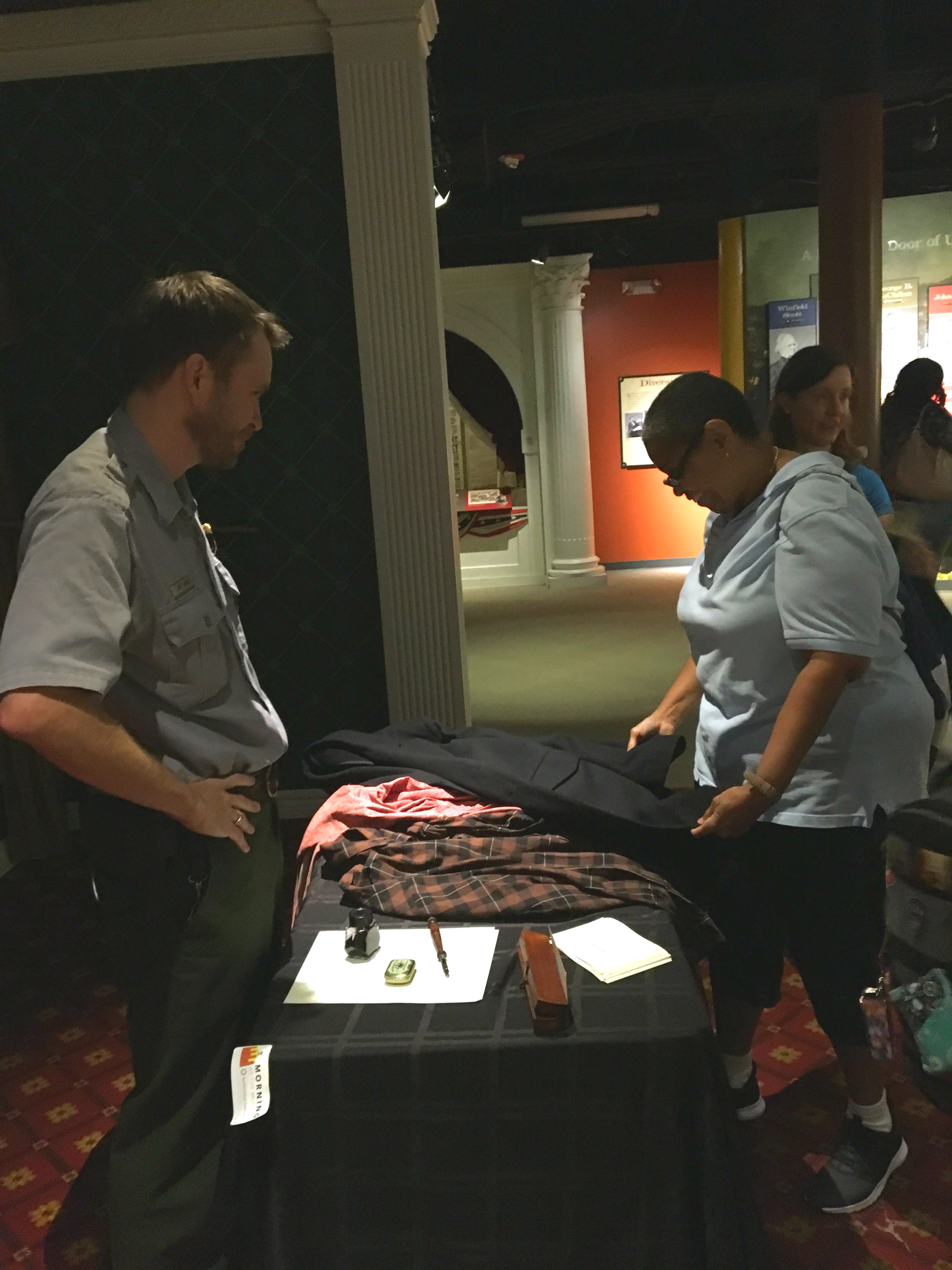 In the Ford's Theatre Museum, a ranger speaks with a visitor. The visitor is touching 1860s period-style clothing that are similar to museum artifacts. Also on the table is an 1860s-style pen and inkwell, and wooden pen box.    