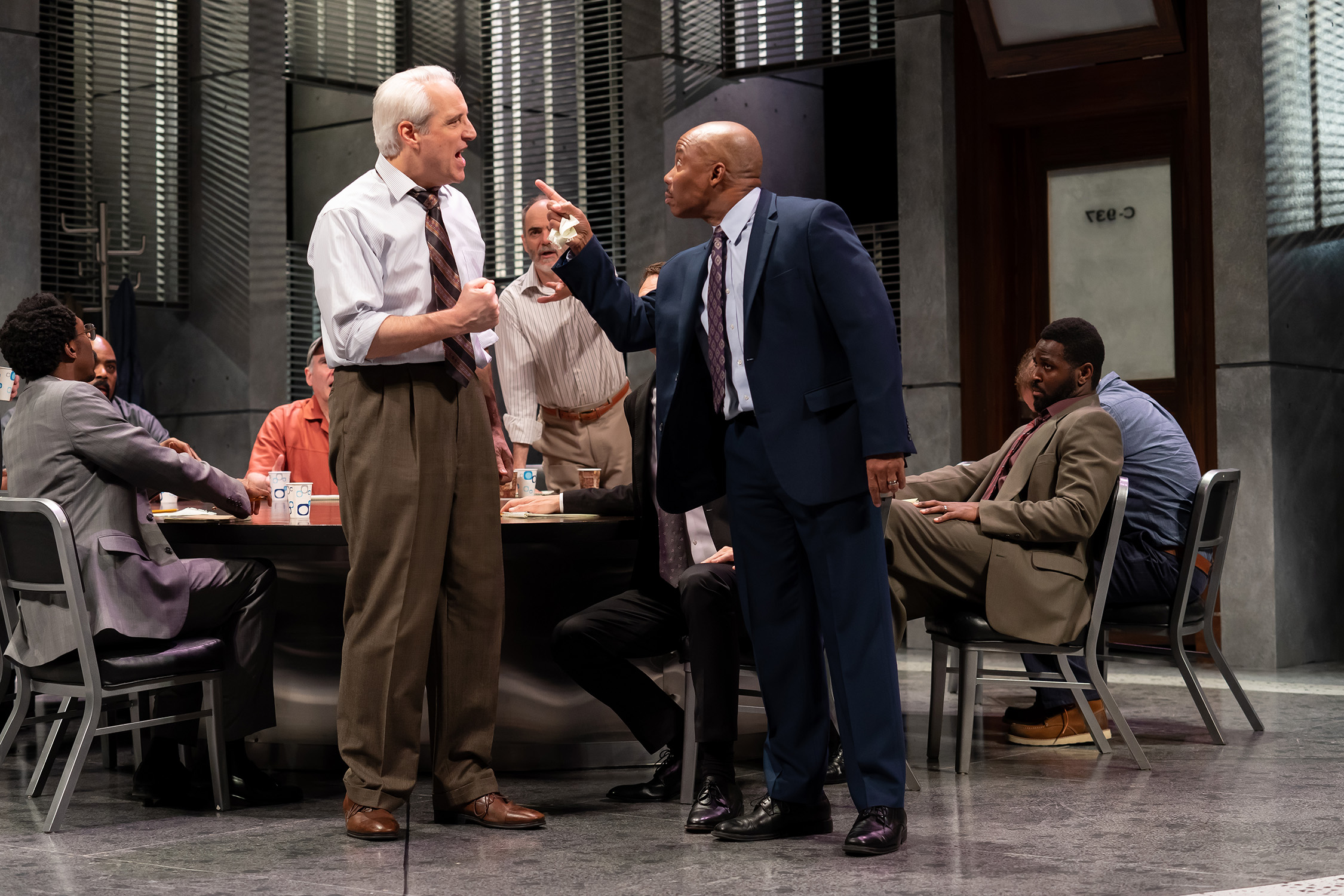 Two jurors (actors Michael Russotto and Erik King) stand, exchanging angry words in a deliberation room. Around them, seven other seated jurors sit with surprised faces. Image is from the Ford’s Theatre 2019 production of Twelve Angry Men, directed by Sheldon Epps. Photo by Scott Suchman