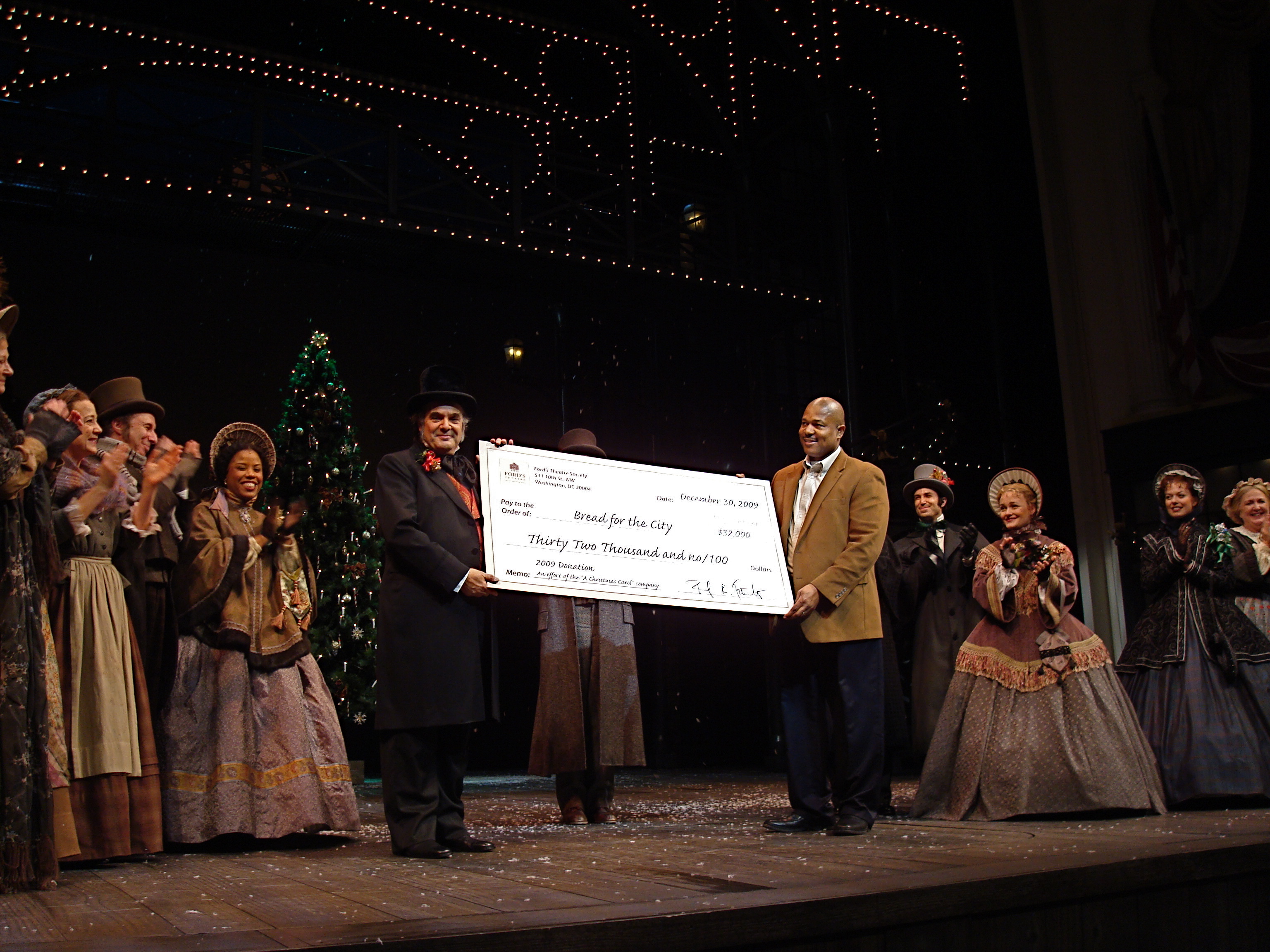 The 2009 cast of "A Christmas Carol" at Ford's Theatre stand in costume on stage. Lead actor Edward Gero is shown presenting a giant check to Bread for the City for $32,000. 