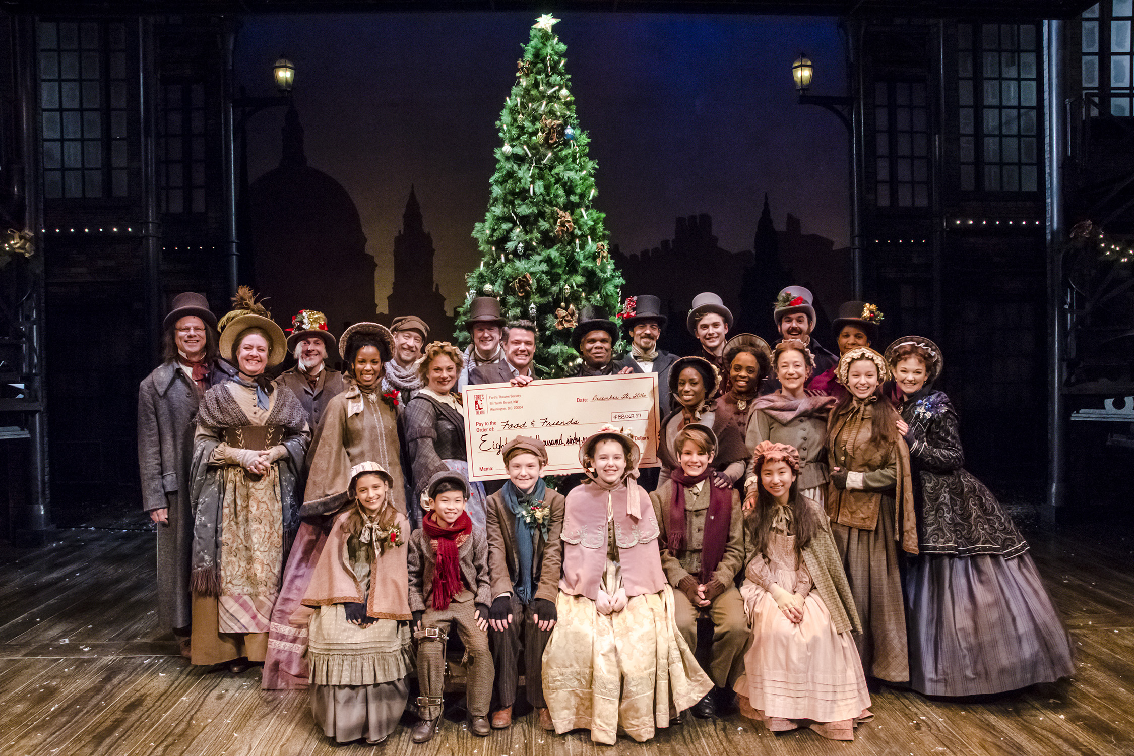 The 2016 cast of "A Christmas Carol" stand on stage in costume with staff from Food & Friends and a giant check for more than $88,000. 