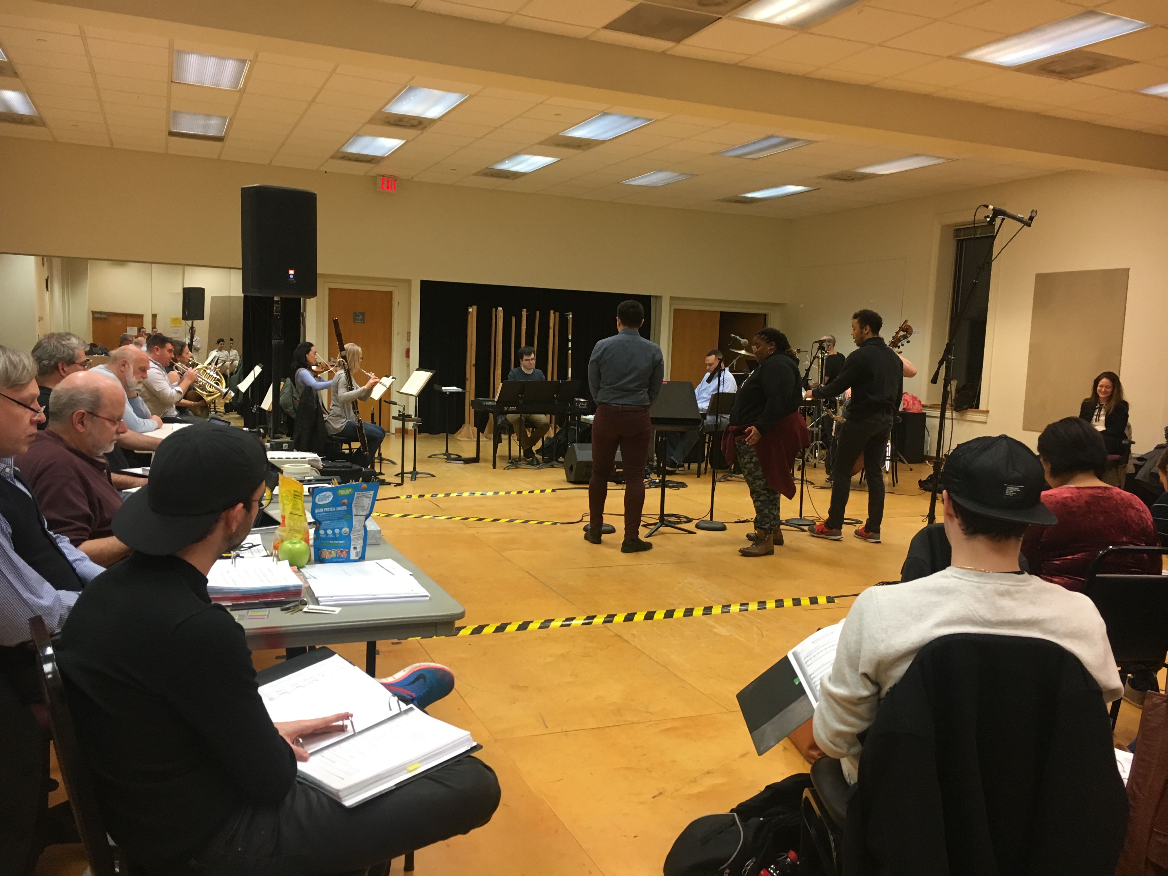 Three actors stand at microphones in the middle of a rehearsal room. A band featuring reed, string and brass instruments, a keyboard and drums is behind them.